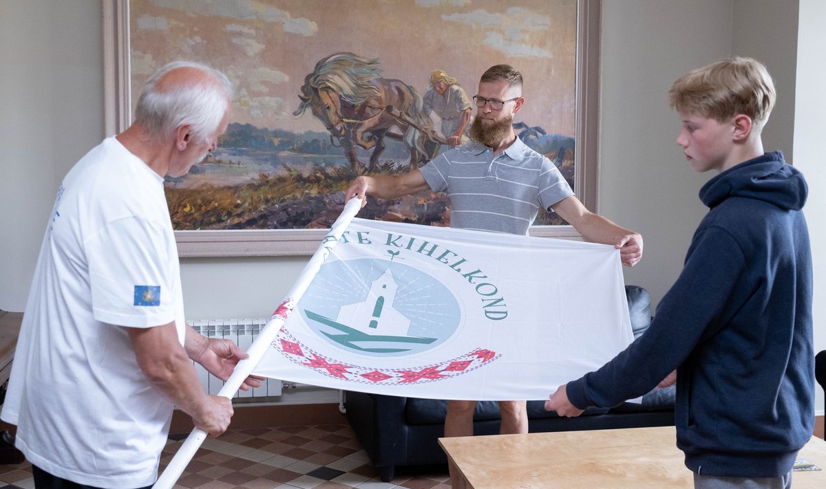 Pärliine pererühma eakamad liikmed pakivad Antslas kihelkonna lippu noorte tantsupeole kaasa võtmiseks ja rongkäigus lehvima panemiseks. Tagas silmab toimetamisi 1950ndasit pärinev kündev Kalevipoeg rahvamaja seinal.