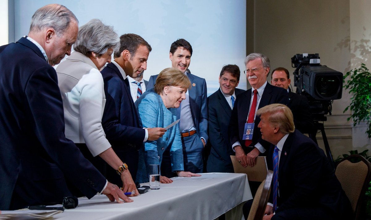 G7 kohtumise samast stseenist on rohkem levinud tõsisem kaader, kuid sel pildil on Prantsuse riigipea Emmanuel Macroni kõrval naeratav Saksa liidukantsler Angela Merkel (helesinises) tema taga Kanada peaminister Justin Trudeau ja kõige parempoolsena USA president Donald Trump. Trumpi kõrval muigab tema riiklik julgeolekunõunik John Bolton.